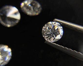 May include: A close-up of several round-cut diamonds, with one held by tweezers. The diamonds are clear and sparkling, reflecting light. The background is black, emphasizing the brilliance of the gemstones.
