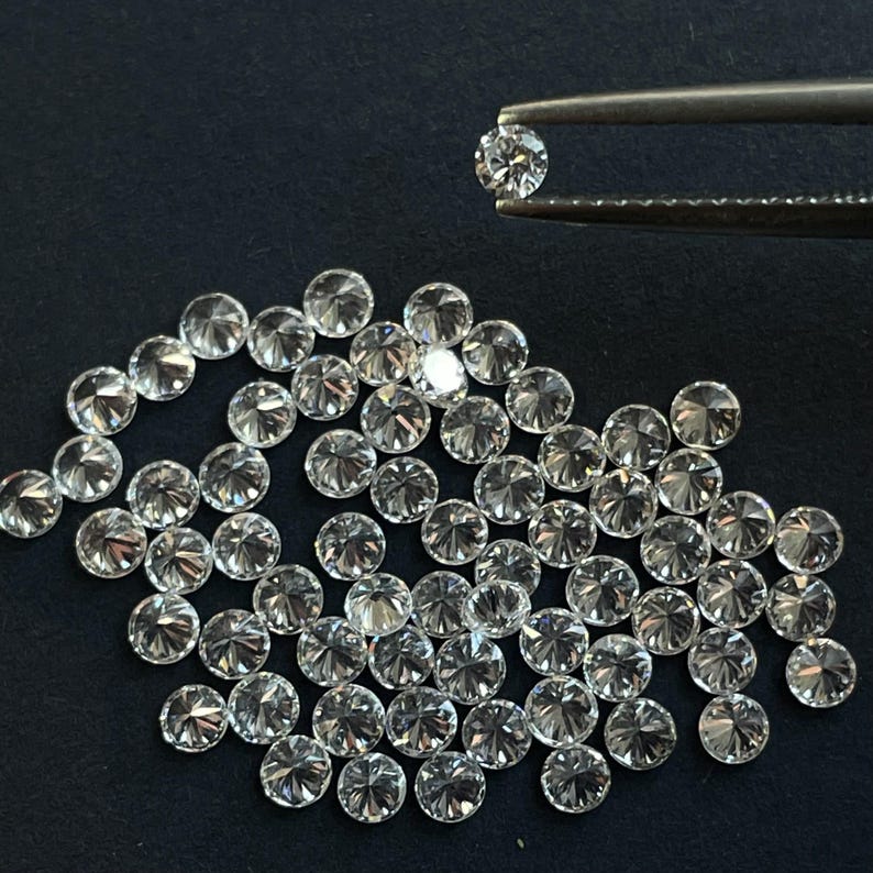 May include: A close-up of a pile of round, clear, faceted diamonds. The diamonds are arranged in a loose, random pattern. One diamond is held in tweezers above the pile.