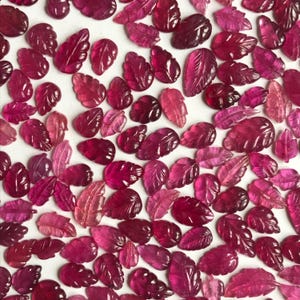 Op de afbeelding: Een verzameling gesneden edelsteenbladeren in verschillende tinten rood en roze. De bladeren zijn ingewikkeld gedetailleerd, met zichtbare aderen en texturen. De edelstenen zijn verspreid over een wit oppervlak.