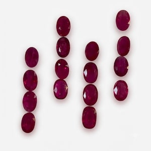 Könnte beinhalten: Vier vertikale Reihen von oval geschliffenen Rubinen, angeordnet vor einem schlichten weißen Hintergrund. Die Edelsteine weisen eine tief gesättigte rote Farbe mit facettierten Oberflächen auf, die das Licht reflektieren und ihre Transparenz betonen.