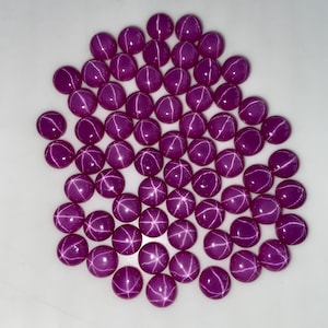 May include: A collection of round, polished, star-shaped gemstones in a deep magenta hue. Each gem displays a six-rayed star pattern across its surface. The gems are arranged closely together against a white background, showcasing their vibrant color and unique optical effect.