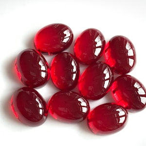 May include: A collection of ten oval-shaped, translucent, ruby-red glass stones. The stones are smooth and polished, reflecting light. They are arranged on a white surface, showcasing their vibrant color and glossy texture. These decorative stones are ideal for crafts.