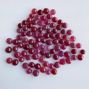 Puede incluir: Un conjunto de pequeñas gemas redondas y pulidas en tonos rojos y morados. Las gemas están dispersas sobre una superficie blanca, con variaciones en tamaño y saturación de color.