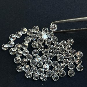 May include: A close-up of a pile of small, round, clear diamonds. The diamonds are arranged in a random pattern and are all about the same size. One diamond is being held by tweezers.