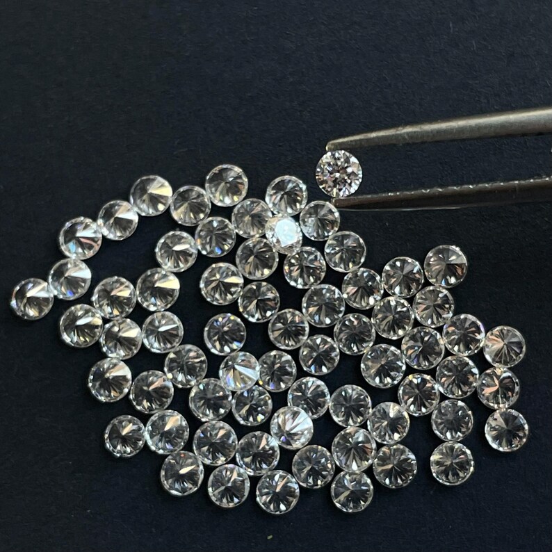 May include: A close-up of a pile of small, round, clear diamonds. The diamonds are arranged in a random pattern and are all about the same size. One diamond is being held by tweezers.