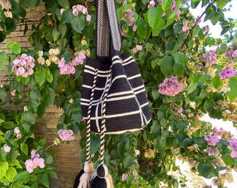 May include: Black and white woven shoulder bag with a geometric pattern. The bag has a long strap and decorative tassels in black, beige, and white. The bag is held up against a backdrop of green foliage and pink flowers.