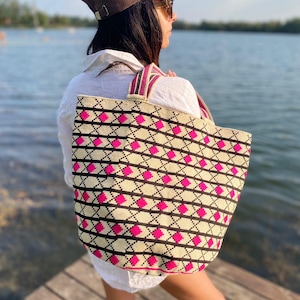 May include: A large, beige tote bag with a black and pink geometric pattern. The bag has thick, pink and brown straps.