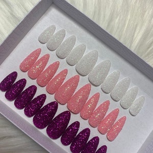 May include: A set of 10 false nails in a white box. The nails are in three colours: white, pink, and purple. Each nail is covered in glitter.