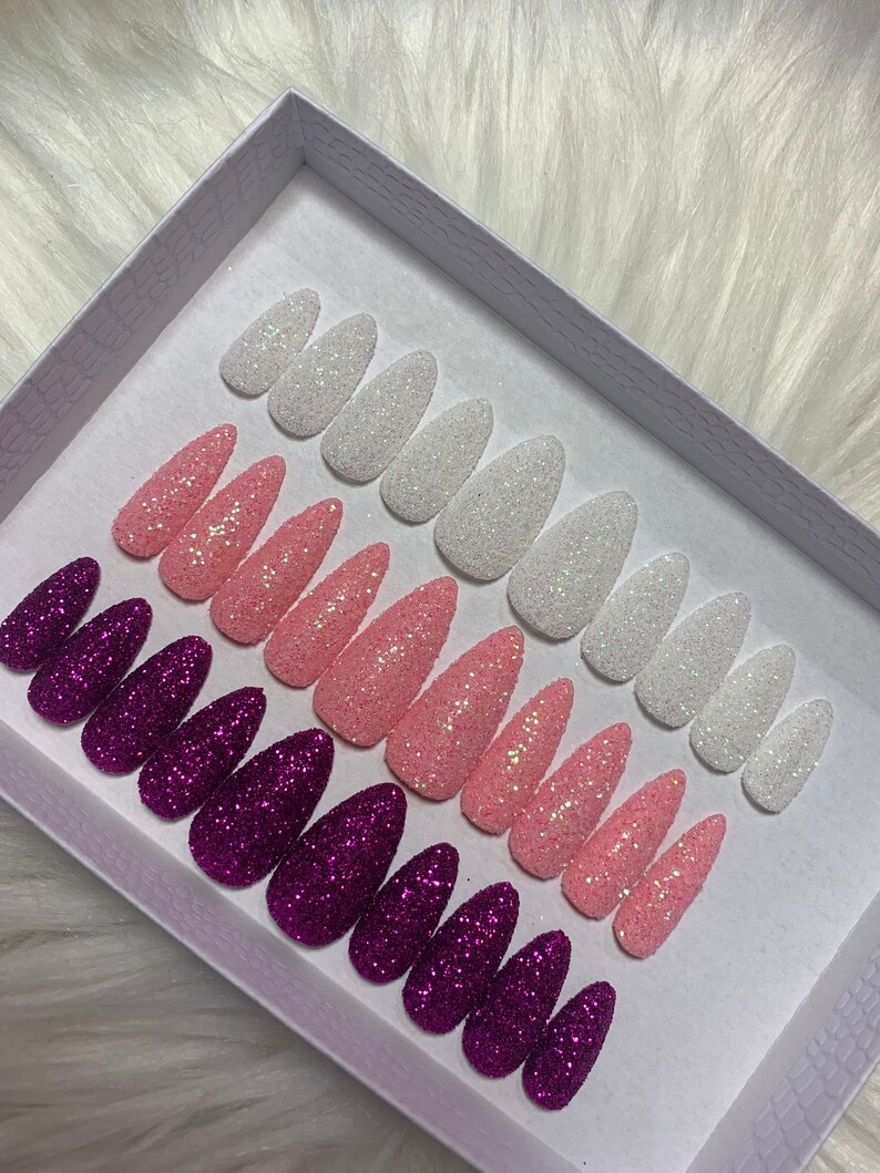 May include: A set of 10 false nails in a white box. The nails are in three colours: white, pink, and purple. Each nail is covered in glitter.