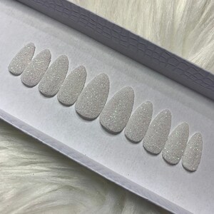 May include: A set of ten white oval-shaped press-on nails with a sparkly glitter finish.