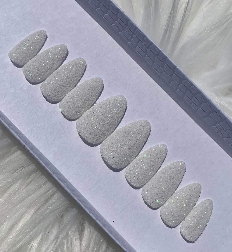 May include: A set of ten white press-on nails with a sparkly finish. The nails are arranged in a row on a white surface.