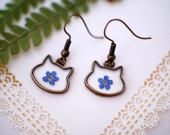 Puede incluir: Pendientes con forma de gato con marco bronce. Cada pendiente tiene un fondo blanco y una pequeña flor azul. Los pendientes cuelgan de ganchos para las orejas de bronce. Un accesorio encantador.