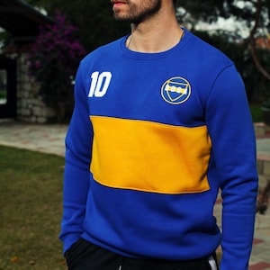 Può includere: Una felpa blu e gialla con il numero 10 bianco e un logo sul davanti. La felpa ha un collo rotondo e maniche lunghe.