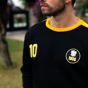 May include: Black sweatshirt with yellow accents, including a yellow collar and shoulder stripe. The number "10" is on the left sleeve, and a circular patch with "1909" is on the chest. The person is wearing a silver chain.