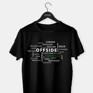 Op de afbeelding: Zwart T-shirt met een wit woordwolkontwerp met de woorden "Buitenspel" en "Voetbal" in groen. De woordwolk bevat andere voetbalgerelateerde termen zoals "wedstrijd", "hoek", "middenvelder", "gele kaart", "coach", "vrije trap", "strafschop", "negentig minuten", "scheidsrechter", "fifa", "uefa", "aftrap", "speeldag", "doelman", "bier", "aanvaller", "jubileum", "schot", "uitwedstrijden", "rode kaart".