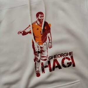 K&ouml;nnte beinhalten: Wei&szlig;er Hoodie mit einer roten und orangenen Grafik eines Fu&szlig;ballspielers mit dem Text "GHEORGHE HAGI" auf der Vorderseite.