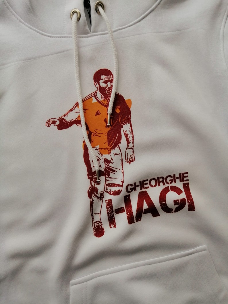 K&ouml;nnte beinhalten: Wei&szlig;er Hoodie mit einer roten und orangenen Grafik eines Fu&szlig;ballspielers mit dem Text "GHEORGHE HAGI" auf der Vorderseite.