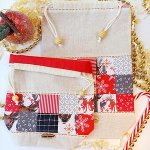Könnte beinhalten: Zwei handgefertigte Stoffgeschenktüten mit roten und weißen Patchwork-Designs. Die kleinere Tasche hat ein Rentier- und Schneeflockenmuster. Die größere Tasche ist schlicht mit rotem Besatz. Beide Taschen haben Holzknöpfe und die kleinere Tasche hat ein "Hand Made"-Etikett.