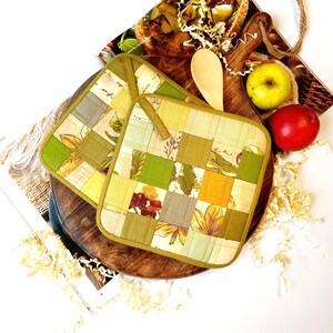 Peut inclure: Deux maniques matelassées vertes et jaunes avec un motif floral. Les maniques sont sur une surface en bois avec une cuillère et des pommes.