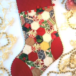 May include: A red and white Christmas stocking with a patchwork design featuring hexagons and snowflakes. The stocking has a red cuff with white pom-poms and a green and white striped trim.