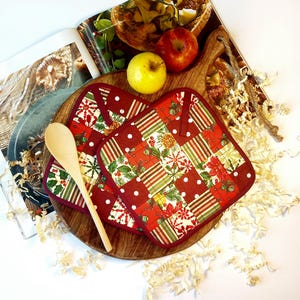 Peut inclure: Une planche à découper en bois avec deux pommes, une cuillère en bois et deux maniques matelassées rouges et blanches sur le thème de Noël. Les maniques ont un motif patchwork avec des flocons de neige, des rayures et des motifs floraux. Un magazine est ouvert en arrière-plan.