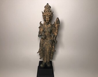 Op de afbeelding: Een bronzen beeld van een godheid, staand op een zwarte basis. De figuur is versierd met ingewikkelde details, waaronder een kroon en sierlijke kleding. Het beeld houdt een voorwerp in één hand en heeft een serene uitdrukking.