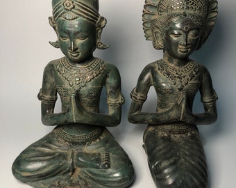 Op de afbeelding: Twee groene bronzen beelden van knielende figuren met gevouwen handen in gebed. De figuren dragen uitgebreide hoofdtooien en traditionele kleding.