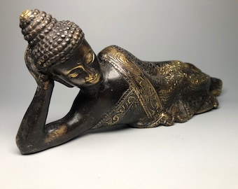 May include: A bronze reclining Buddha statue with gold accents. The statue is lying on its side with its head resting on its hand. The Buddha is wearing a robe with intricate details.