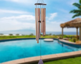 May include: A wind chime with a dark brown wooden top and six metal tubes. The tubes are a light copper color and have a striped pattern. The wind chime is hanging from a black string and has a dark brown wooden clapper at the bottom. The wind chime is hanging in front of a swimming pool and a palm tree.