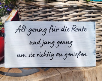 May include: A rectangular wooden sign with white paint and black lettering. The text reads "Alt genug für die Rente und jung genug um sie richtig zu genießen."
