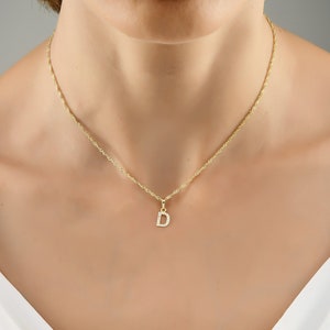 May include: A gold necklace featuring a pendant in the shape of the letter "D", adorned with small crystals. The necklace has a fine chain. The jewellery is displayed on a person's neck.