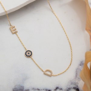Op de afbeelding: Gouden ketting met een blauwe boze oog bedel en twee letterbedels, E en D, bezet met heldere zirkonia stenen.