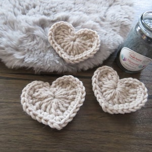May include: Three crocheted hearts in a light beige colour. The hearts are on a wooden surface.