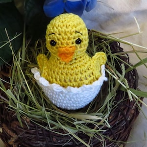 Könnte beinhalten: Ein gehäkeltes gelbes Küken in einer weißen gehäkelten Eierschale, das in einem braunen Weidenkorb mit getrocknetem Gras sitzt.