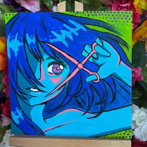 May include: A vibrant pop art painting on canvas depicts a stylized figure with blue hair and skin, holding pink scissors. The figure's eye is purple, and the background is a mix of green and blue. The artwork is displayed on a small wooden easel.