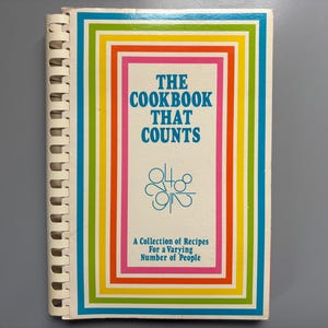 Op de afbeelding: Een spiraalgebonden kookboek getiteld "The Cookbook That Counts" met een kleurrijke rechthoekige rand. De omslag bevat blauwe tekst en een verzameling recepten voor een wisselend aantal personen.