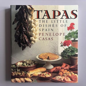 May include: A cookbook titled "TAPAS" with the subtitle "The Little Dishes of Spain" by Penelope Casas. The cover features a variety of Spanish tapas dishes, including a tortilla, sausages, and olives, along with red flowers and dried peppers.