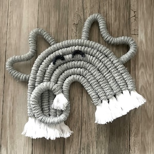 May include: A gray macrame rainbow wall hanging with white tassels. The rainbow has a whimsical face with black eyes.