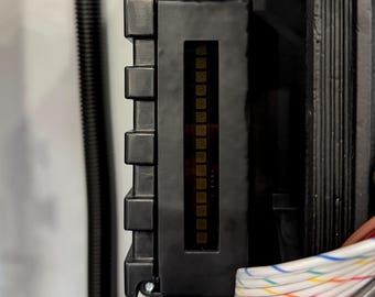 May include: A black rectangular electronic component housing featuring a vertical display window with a series of small square indicators. The unit is connected to a bundle of multi-colored wires and a black corrugated cable, with a yellow wire visible.