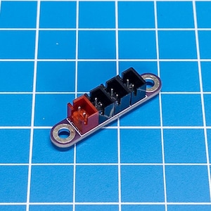 May include: A small electronic component with three black and one red connector ports. The component is mounted on a purple base with two circular mounting holes. The background is a blue grid.