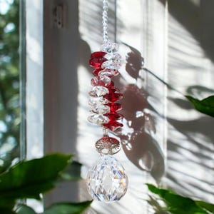 Könnte beinhalten: Ein Kristall-Sonnenfänger mit roten und klaren Glasperlen, der an einem Fenster hängt. Der Sonnenfänger wirft Regenbögen an die Wand.