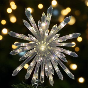 May include: A clear, starburst-shaped Christmas tree topper with multiple crystal-like points. The topper is illuminated with internal lights, creating a sparkling effect. The background features blurred, warm-toned lights.