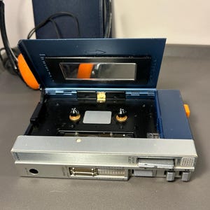 May include: A blue and silver portable cassette player with the cassette compartment lid open. The device features a sliding volume control, a stop/eject button, and a microphone port. The Sony brand name is printed on the silver front panel.