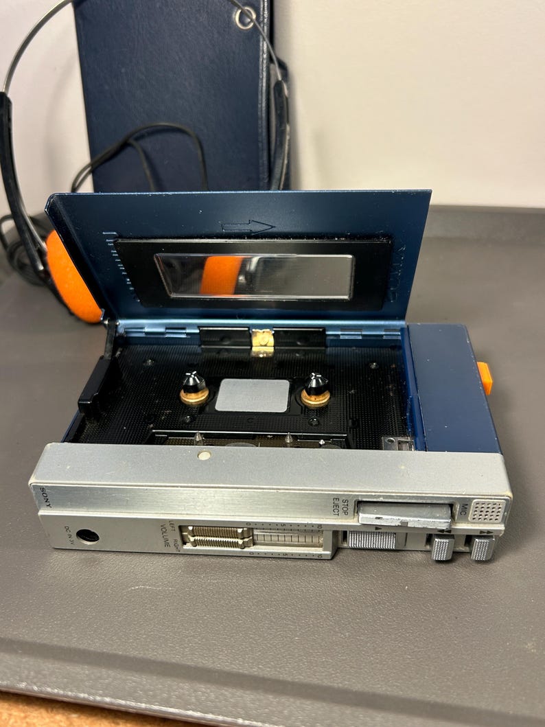 May include: A blue and silver portable cassette player with the cassette compartment lid open. The device features a sliding volume control, a stop/eject button, and a microphone port. The Sony brand name is printed on the silver front panel.