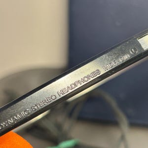 May include: A black plastic headband component of headphones featuring the embossed white text DYNAMIC STEREO HEADPHONES MDR-010 and a circular L symbol indicating the left side. The surface is smooth and reflective with a visible orange foam piece.