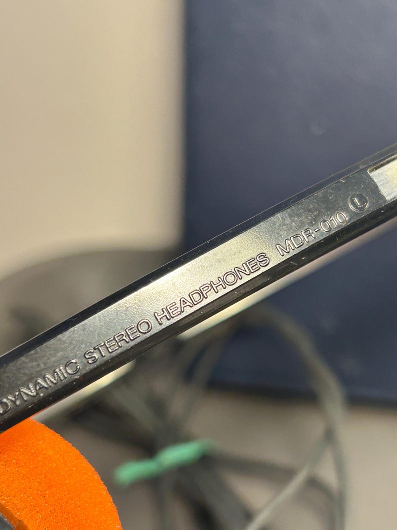 May include: A black plastic headband component of headphones featuring the embossed white text DYNAMIC STEREO HEADPHONES MDR-010 and a circular L symbol indicating the left side. The surface is smooth and reflective with a visible orange foam piece.