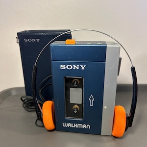 May include: A blue and silver Sony Walkman cassette player with a clear window, an orange button, and a white arrow graphic. The device features the Sony and Walkman logos. Included are black wired headphones with bright orange foam ear pads.