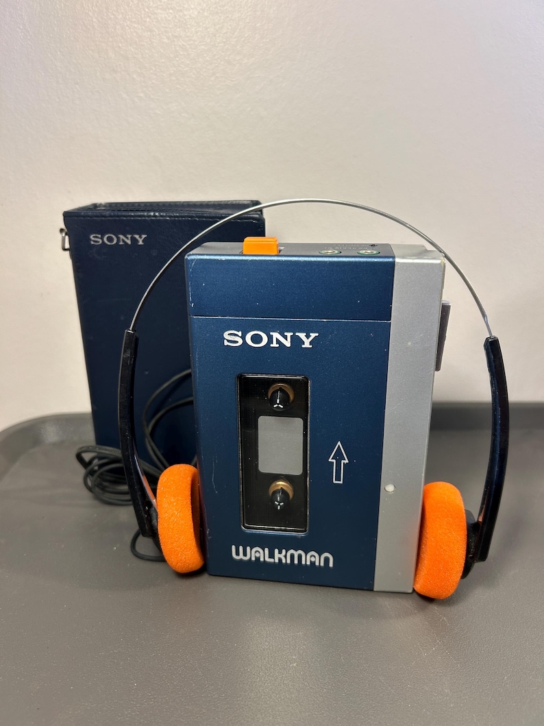 May include: A blue and silver Sony Walkman cassette player with a clear window, an orange button, and a white arrow graphic. The device features the Sony and Walkman logos. Included are black wired headphones with bright orange foam ear pads.