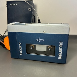 May include: A blue and silver Sony Walkman cassette player with a rectangular viewing window, a white arrow icon, and the Sony Walkman logo printed on the front. A dark blue leather-like carrying case with the Sony logo is positioned behind it.