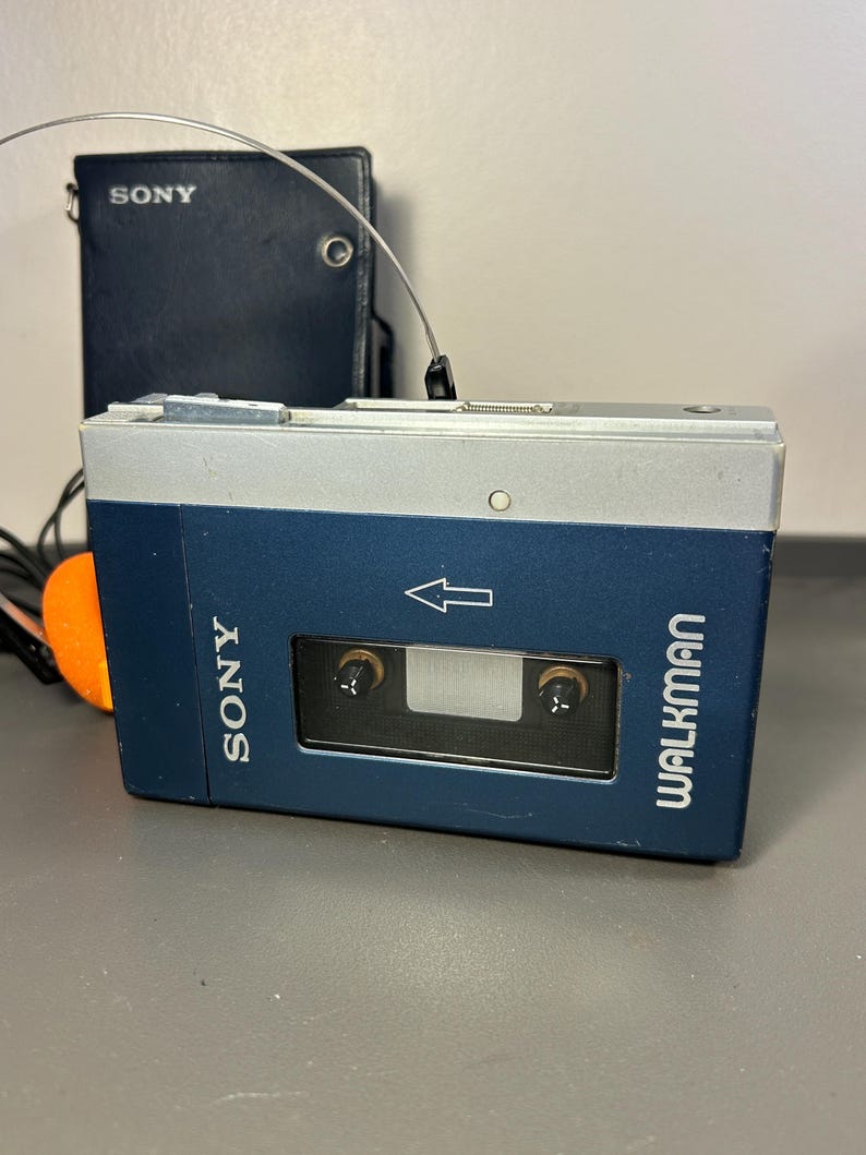 May include: A blue and silver Sony Walkman cassette player with a rectangular viewing window, a white arrow icon, and the Sony Walkman logo printed on the front. A dark blue leather-like carrying case with the Sony logo is positioned behind it.